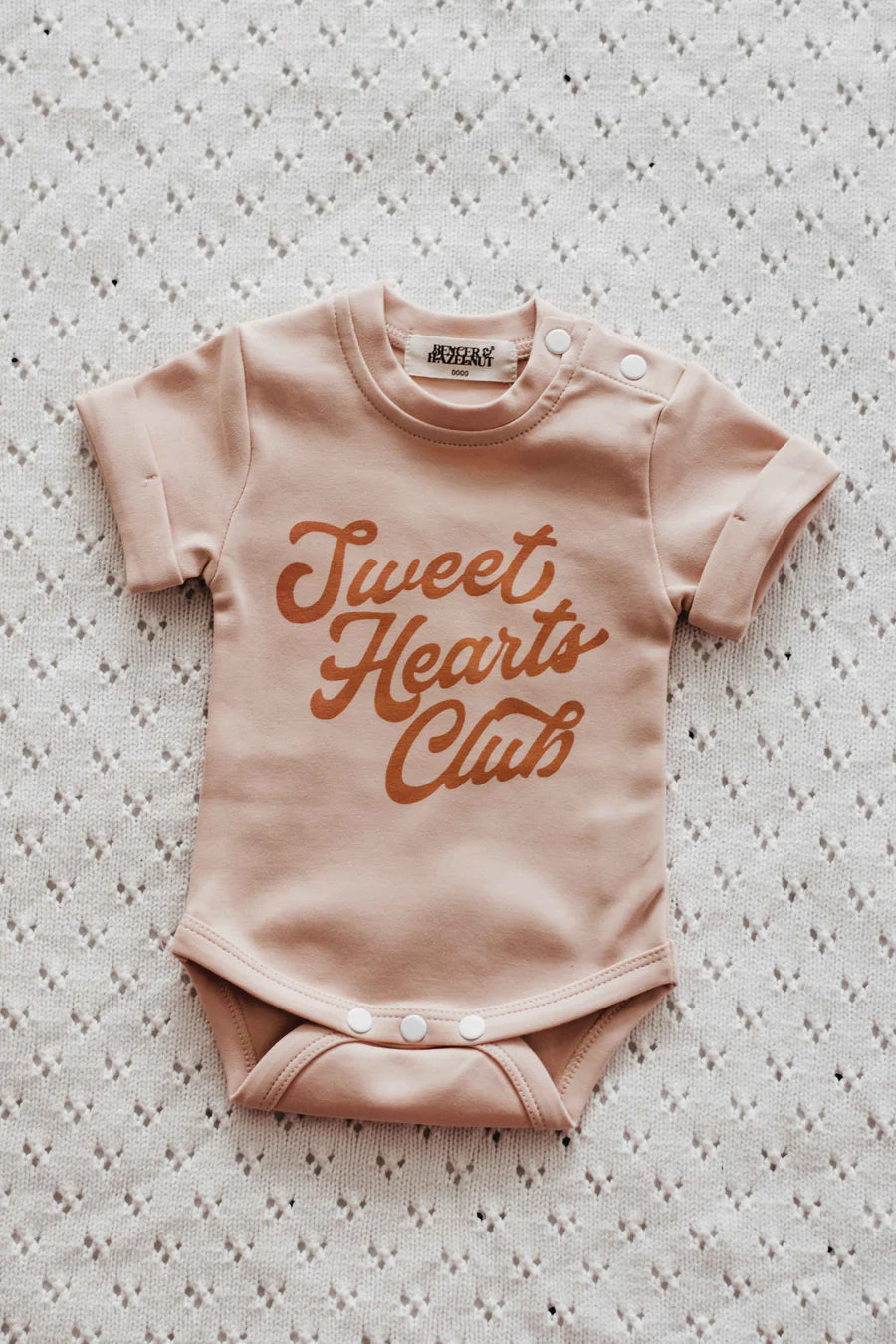 A short-sleeve baby bodysuit in a soft blush pink color, featuring the phrase 'Sweet Hearts Club' printed in a warm terracotta brown script across the chest. The bodysuit is laid flat on a textured, off-white knit blanket, with three snap buttons visible at the crotch and shoulder seams.