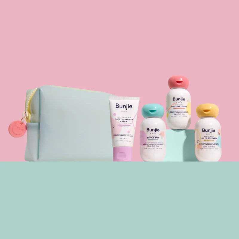 A pastel-colored Bunje baby gift set featuring a light blue zippered pouch, a pink tube, a teal bottle, a white bottle with a pink cap, and a white bottle with a yellow cap, arranged on a split pink and mint green background.