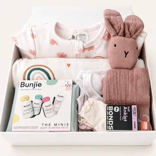 A baby starter pack gift box from Bunjie, containing a light pink bunny-themed bodysuit, a white raincloud blanket, a dusty pink muslin bunny plush, a muslin cloth, a Bond's muslin swaddle, and a Bunjie baby care booklet.