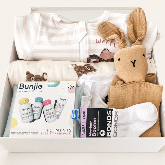 A baby starter pack gift box containing a striped bodysuit, a bunny plush toy, a muslin blanket, a pack of white socks, a boxed set of baby bottles, and a pack of baby wipes, all arranged in a white box.