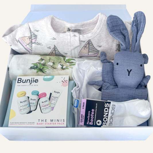 A baby starter pack gift box containing a white long-sleeve baby top with a sailboat print, a plush blue bunny toy, a tube of Bunjie baby skincare, and a pack of Bonds baby booties, all arranged in a light blue box.