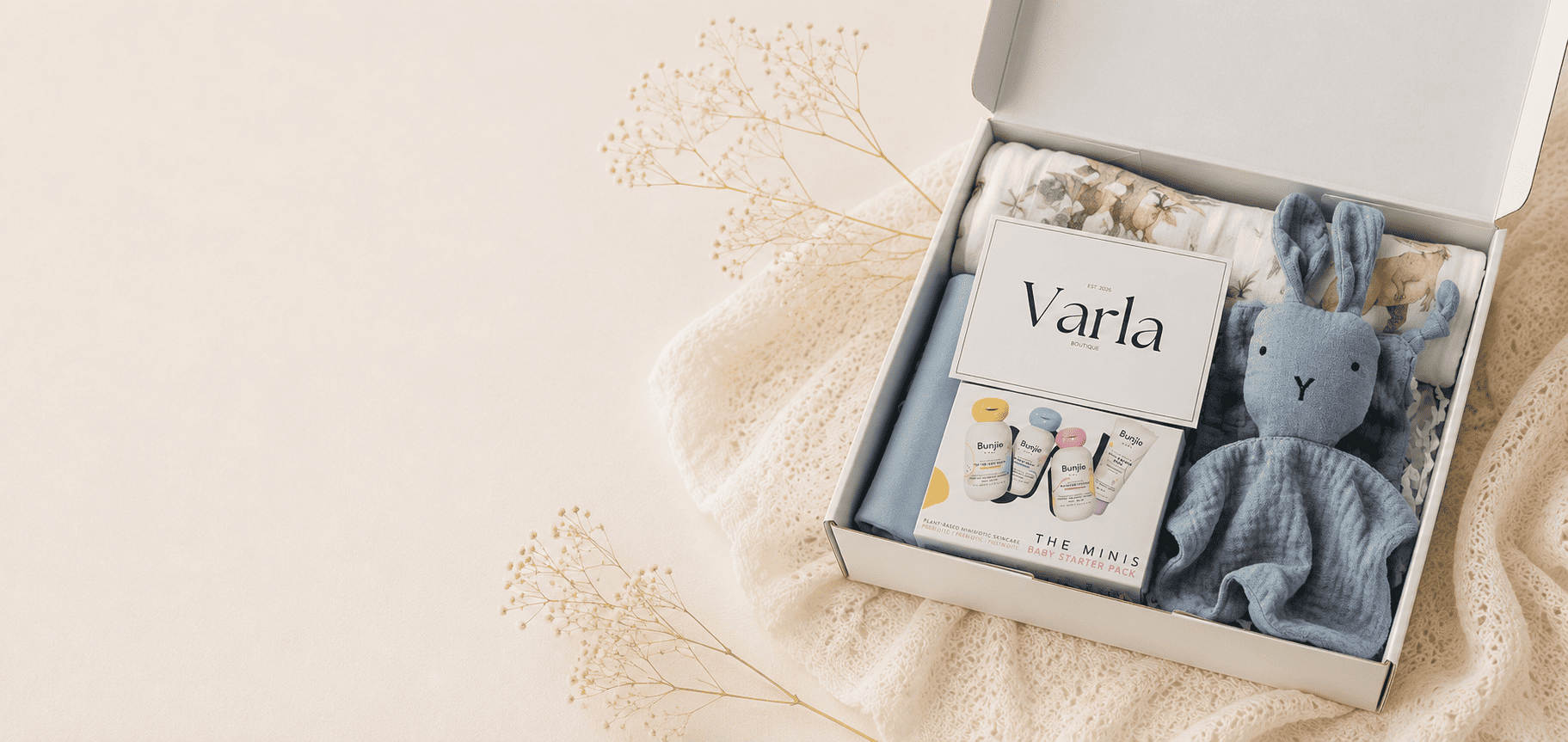 A personalized baby gift box from The Mini Boutique, featuring a blue plush bunny snuggler, a printed card personalized with the name 'Varla', and folded baby clothes inside a white box, styled on a cream lace cloth with dried floral accents.