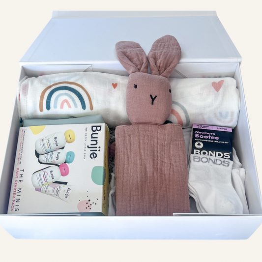 A curated baby gift set arranged neatly inside an open white box. The set includes a dusty rose bunny plush toy, a white muslin blanket with a rainbow and heart print, a box of Bunkite baby formula, a pack of white socks, and a package of Bonds Bond's Bootie socks.