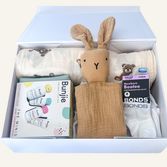 A baby gift set arranged inside an open white box on a light beige background. The set includes a tan muslin bunny-shaped comforter, white muslin swaddles with teddy bear prints, a package of Bond's Newborn Bootie socks, and a package of The Minis Bunjie baby wipes.