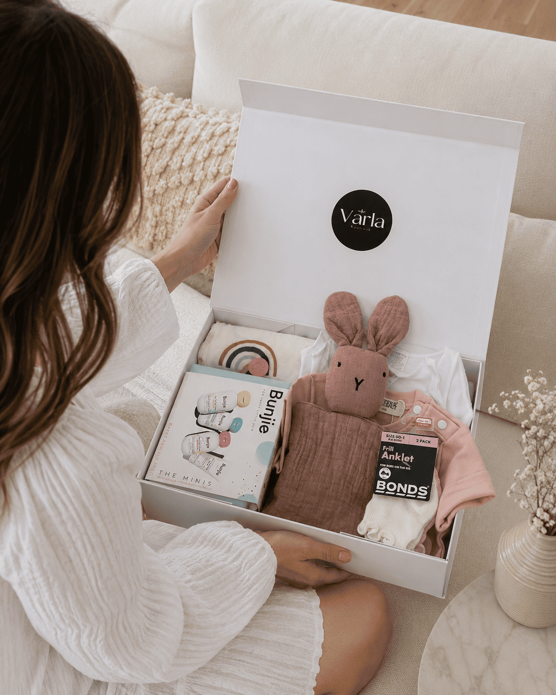 A white gift box opened by a woman in a white ribbed robe, containing baby items including a pink bunny plush toy, a pink blanket, a book, and a small basket. The box lid has a black circular logo reading 'the Varia'.